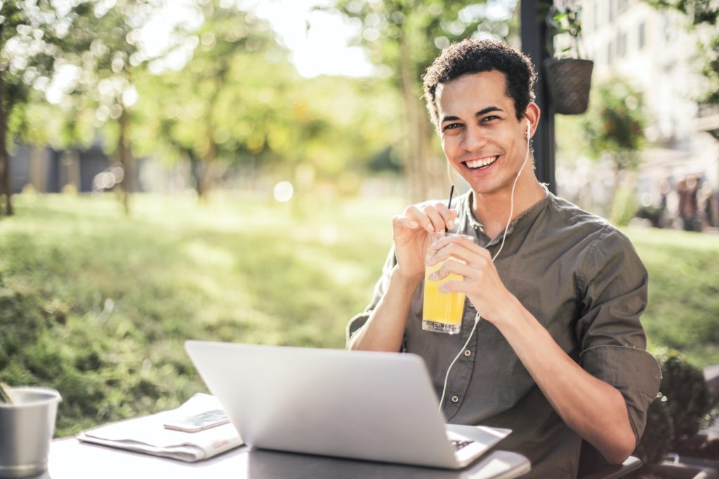 Photo d'un jeune homme souriant devant son ordinateur avec des écouteurs et en train de siroter un jus de fruit
